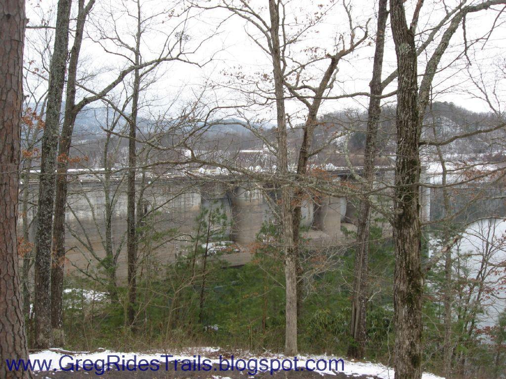 A view of a dam partially obscured by bare trees, with a snowy landscape in the foreground and distant hills in the background. The image captures a serene winter scene. Fontana Village mountain bike trail.