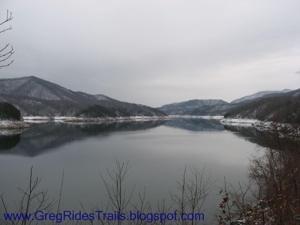 A serene view of a lake surrounded by snow-dusted mountains under a gray sky. The calm water reflects the landscape, creating a tranquil atmosphere. Sparse trees in the foreground add a touch of nature to the scene. Fontana Village mountain bike trail.