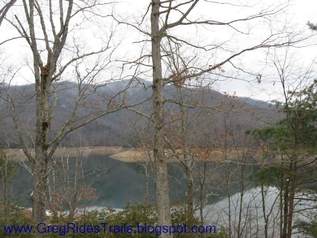 A scenic view of a tranquil lake surrounded by barren trees in a mountainous area. The landscape features a calm reflection on the water and distant hills, creating a peaceful atmosphere. Fontana Village mountain bike trail.