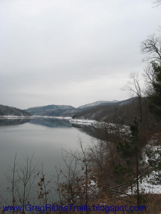 A scenic view of a tranquil lake surrounded by snow-covered hills and trees under a cloudy sky. The still water reflects the landscape, creating a calm and serene atmosphere. Fontana Village mountain bike trail.