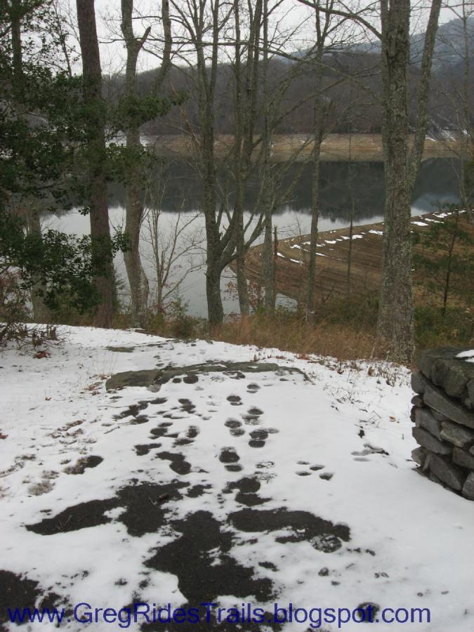 A snowy pathway leading down to a calm body of water, surrounded by bare trees and a hillside in the background. Footprints are visible in the snow, indicating previous activity. Fontana Village mountain bike trail.