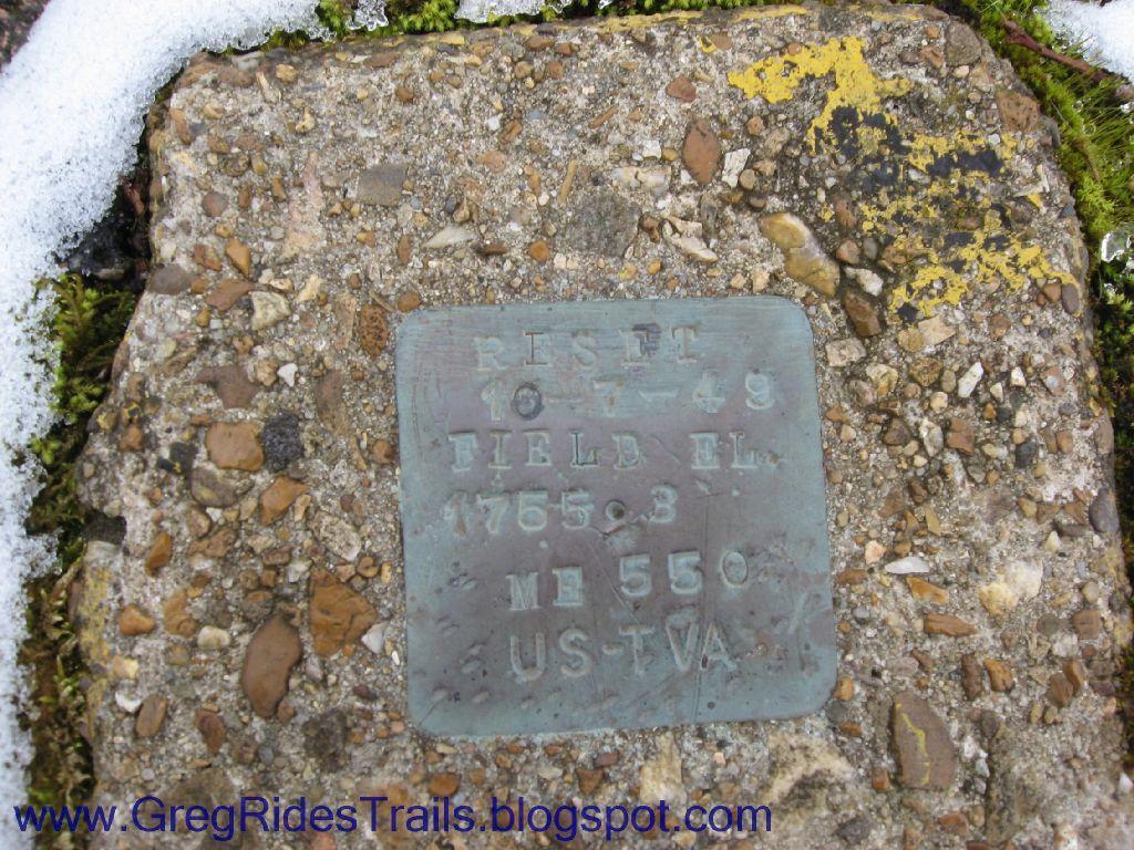 A close-up view of a metal survey marker set into a stone surface, featuring engraved text that reads "RESET 10-17-49 FIELD EL. 1765.3 ME 550 UST-VA." The marker is surrounded by gravel and patches of snow, indicating a natural outdoor setting. Fontana Village mountain bike trail.