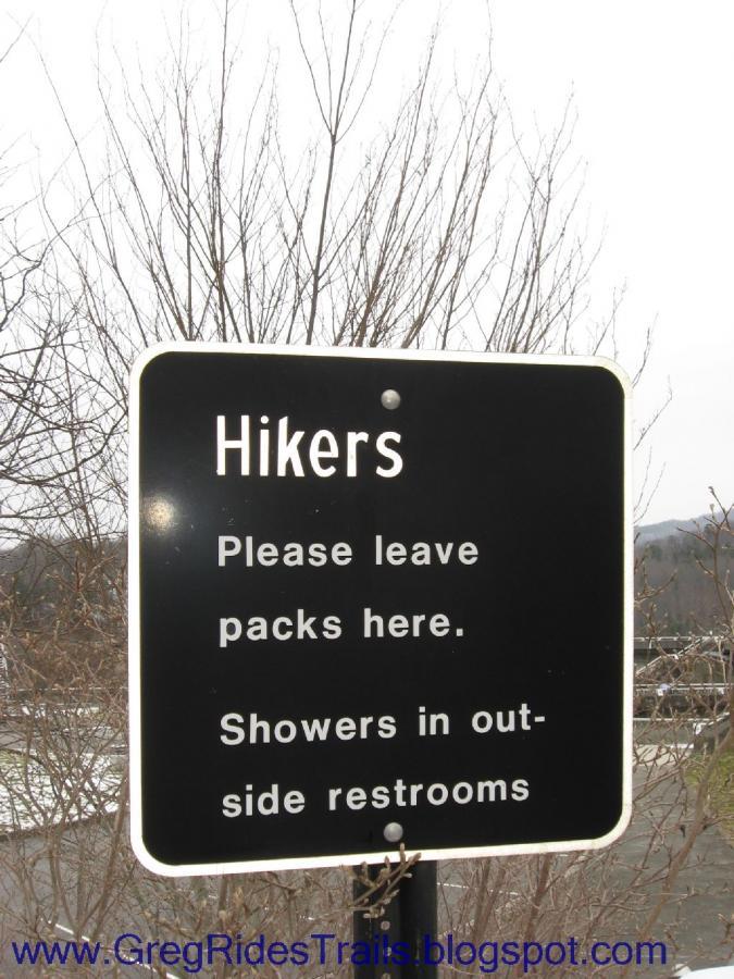 A black and white sign instructing hikers to leave their packs in a designated area. The sign reads: "Hikers. Please leave packs here. Showers in outside restrooms." There are bare trees in the background, indicating a possibly cool or early spring setting. Fontana Village mountain bike trail.