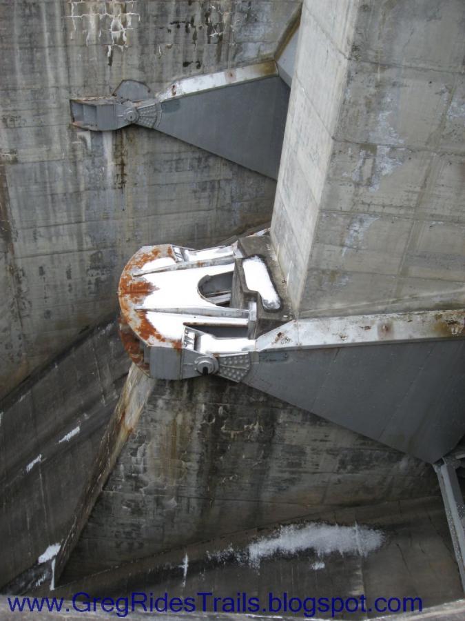 Close-up view of a large, weathered metal structure attached to a concrete wall. The structure features rust and is partially covered in snow, with details of bolts and surface texture visible. The background displays rough, concrete surfaces, suggesting an industrial setting. Fontana Village mountain bike trail.