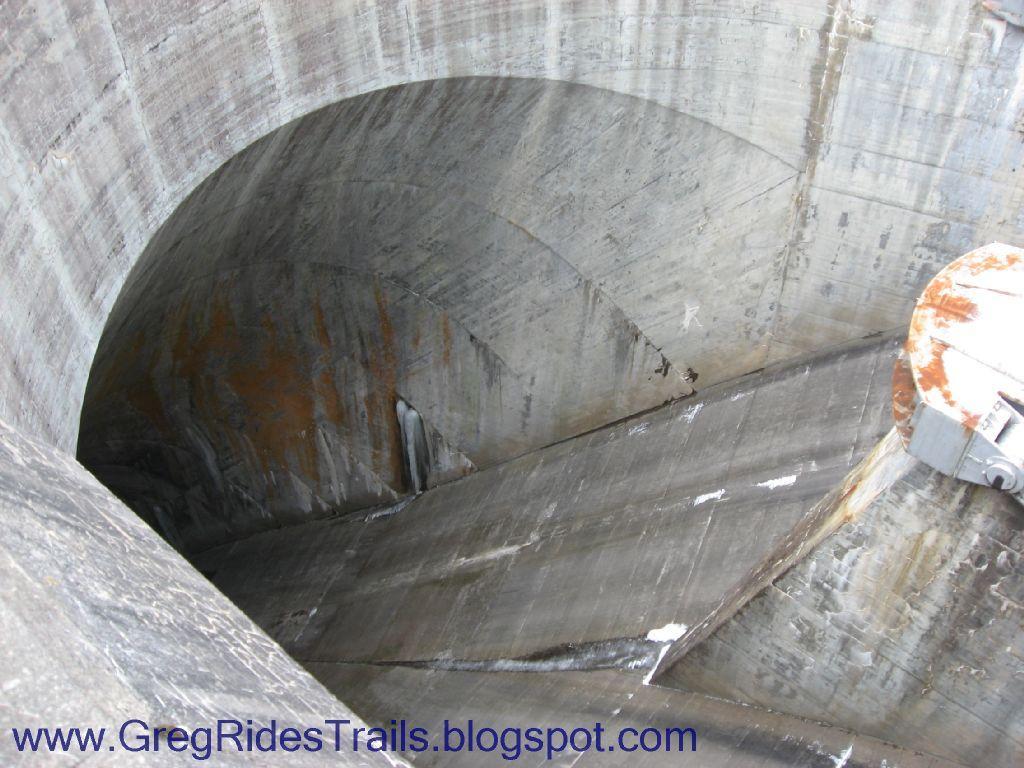 A close-up view of a large concrete structure, likely part of a dam or water reservoir, featuring a curved interior surface with visible wear and rust. The image captures the geometric shapes and lines formed by the structure's construction and the sloped paths leading downward. Fontana Village mountain bike trail.