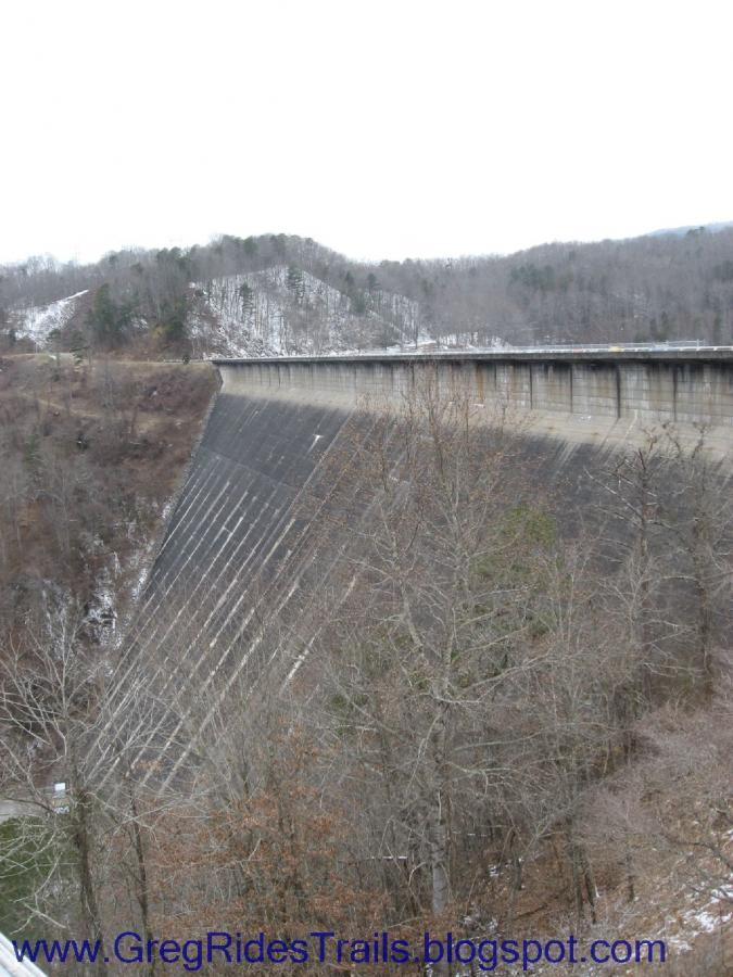 A view of a large concrete dam stretching across a landscape with bare trees, against a backdrop of rolling hills and a cloudy sky. The dam's sloped face and vertical lines are prominent, indicating its structure and purpose. Fontana Village mountain bike trail.