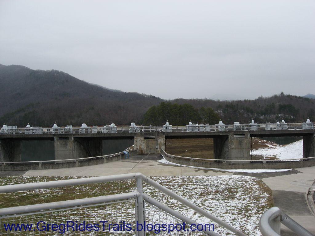 A view of a dam with a concrete structure spanning across a body of water, surrounded by bare trees and mountains in the background. The sky is overcast, and there's a hint of snow on the ground. A walkway leading to the dam is visible in the foreground, along with railings. Fontana Village mountain bike trail.