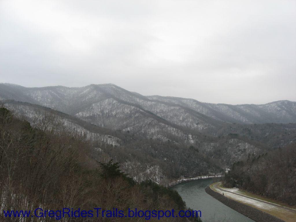 A scenic view of mountainous terrain covered with a light dusting of snow under a cloudy sky. A winding river flows through the valley, bordered by tree-covered slopes. The atmosphere appears calm and serene, capturing the beauty of a winter landscape. Fontana Village mountain bike trail.