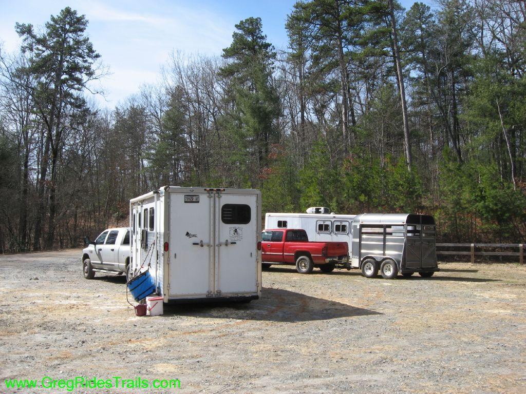 A gravel parking area surrounded by trees, featuring two horse trailers and a pickup truck. One trailer is white with a ramp, and the other is a silver stock trailer. An additional black truck is parked nearby. The scene is set in a wooded area, indicating a potential outdoor or equestrian setting. Jake Mountain Trails mountain bike trail.