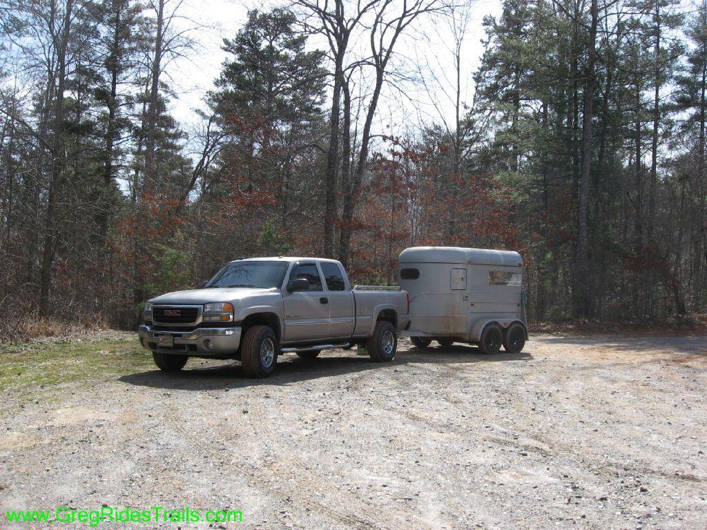 A gray GMC truck parked on a gravel road, towing a silver horse trailer. The scene is surrounded by bare trees and sparse foliage, indicating early spring. Jake Mountain Trails mountain bike trail.