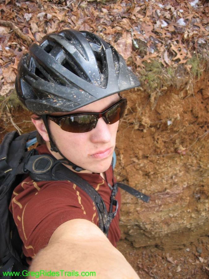 A person wearing a black helmet and sunglasses takes a selfie in a natural setting, with dirt and leaves in the background. The individual has a backpack and is dressed in a brown shirt. The scene suggests an outdoor adventure, likely related to biking or hiking. Jake Mountain Trails mountain bike trail.