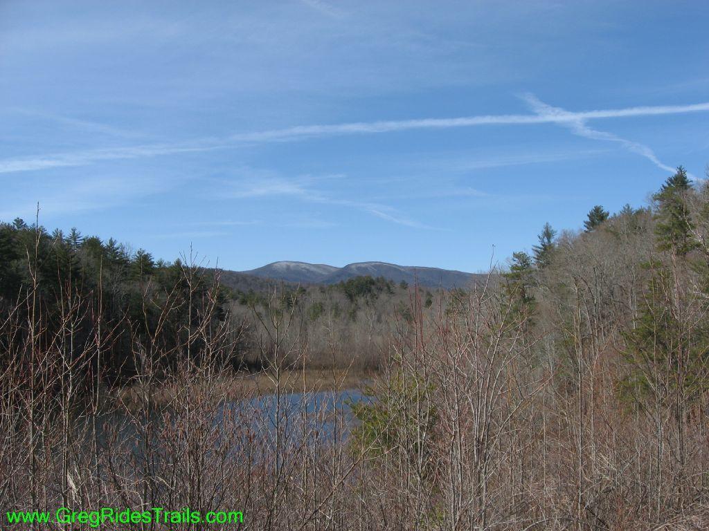 A tranquil landscape view featuring a serene body of water surrounded by bare trees, with rolling mountains in the background under a clear blue sky. Jones Creek Ridge Trail mountain bike trail.