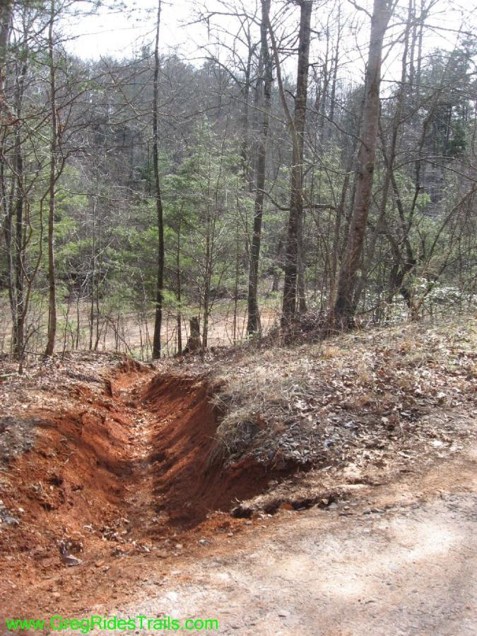 A dirt trail with a deep, narrow trench running alongside it, surrounded by trees and underbrush. The soil appears reddish and dry, with some scattered leaves and rocks. In the background, a wooded area is visible, hinting at a natural landscape. Jake to Bull Mountain Connecter mountain bike trail.