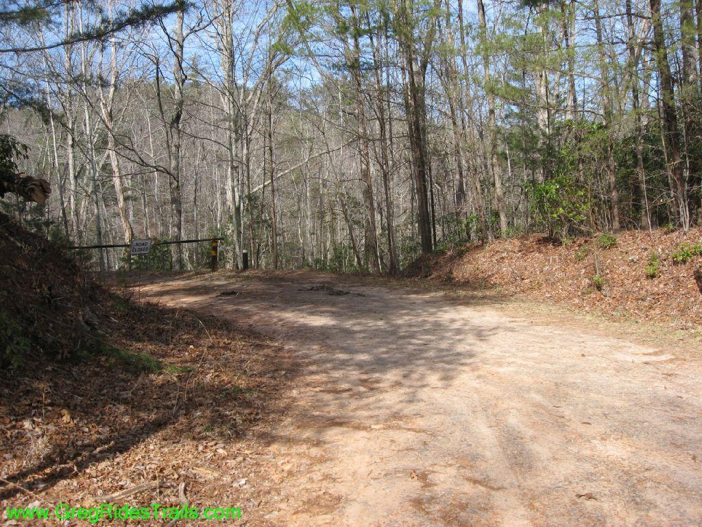 A dirt road winding through a forested area, with leafless trees and sparse greenery visible. A closed gate is positioned at the entrance of the road, with a "Road Closed" sign clearly visible. The scene depicts a tranquil, wooded landscape under a clear blue sky. Jake to Bull Mountain Connecter mountain bike trail.