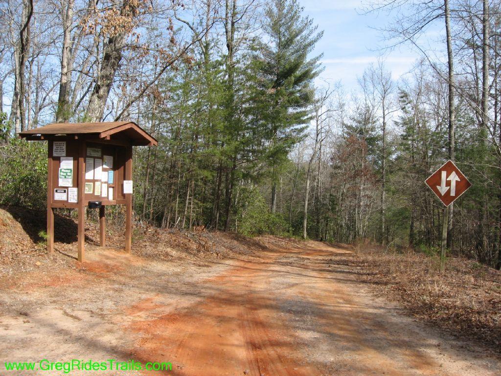 A dirt trail leading into a wooded area, featuring a wooden information kiosk on the left with various trail signs and maps. A brown directional sign indicating a change in trail direction is visible on the right. Tall trees line the path, with a mix of green pines and bare deciduous trees. The sky is partly cloudy. Jake to Bull Mountain Connecter mountain bike trail.