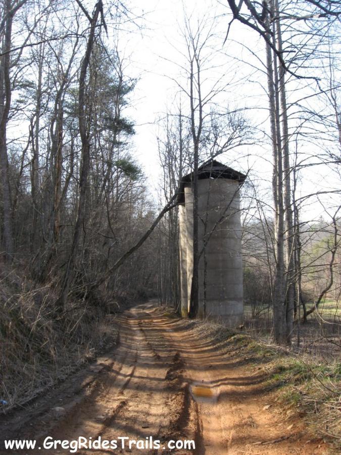 A dirt path winds through a forest, flanked by bare trees, leading toward a tall, cylindrical concrete structure with a flat roof. The scene is set on a clear day, showcasing the natural surroundings and the silhouette of the silo against the sky. Canada Creek mountain bike trail.