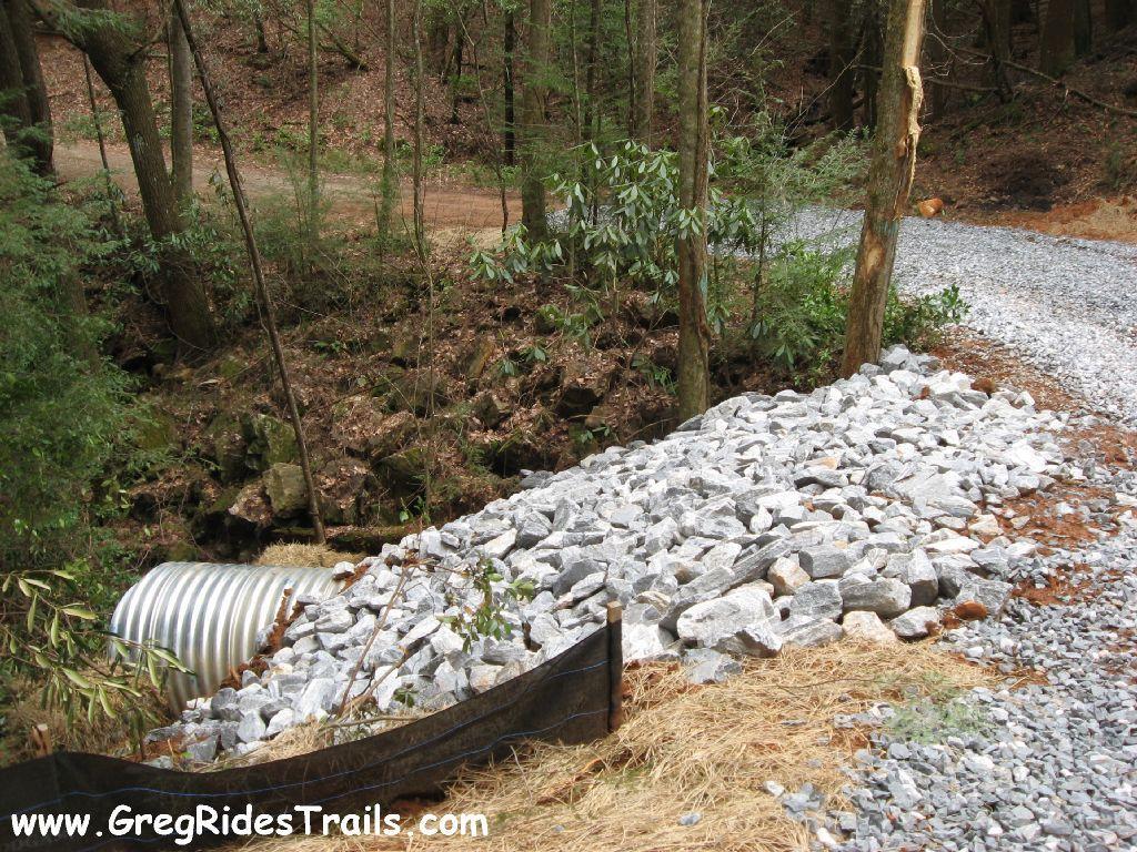 A gravel pathway runs alongside a rocky area with a metal culvert. The foreground features a pile of large, gray stones used for erosion control, while lush greenery and trees line the background. A dirt road can be seen curving through the scene. Montgomery Creek Trail mountain bike trail.
