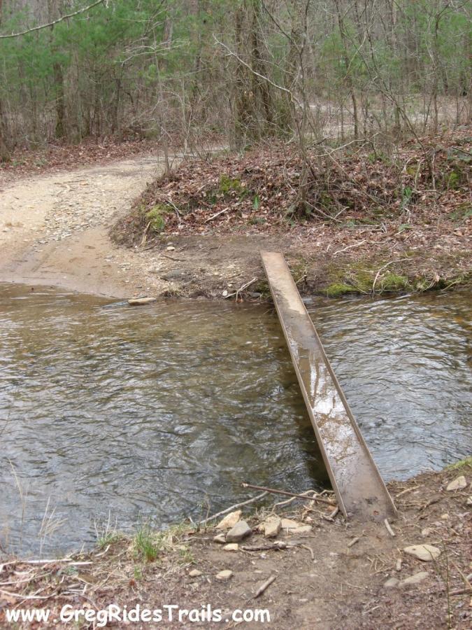 A narrow wooden bridge crossing over a small stream in a forested area. The surrounding landscape features a mix of dirt paths and trees, with some foliage on the ground. The atmosphere is peaceful and natural, characteristic of a hiking or biking trail. Montgomery Creek Trail mountain bike trail.