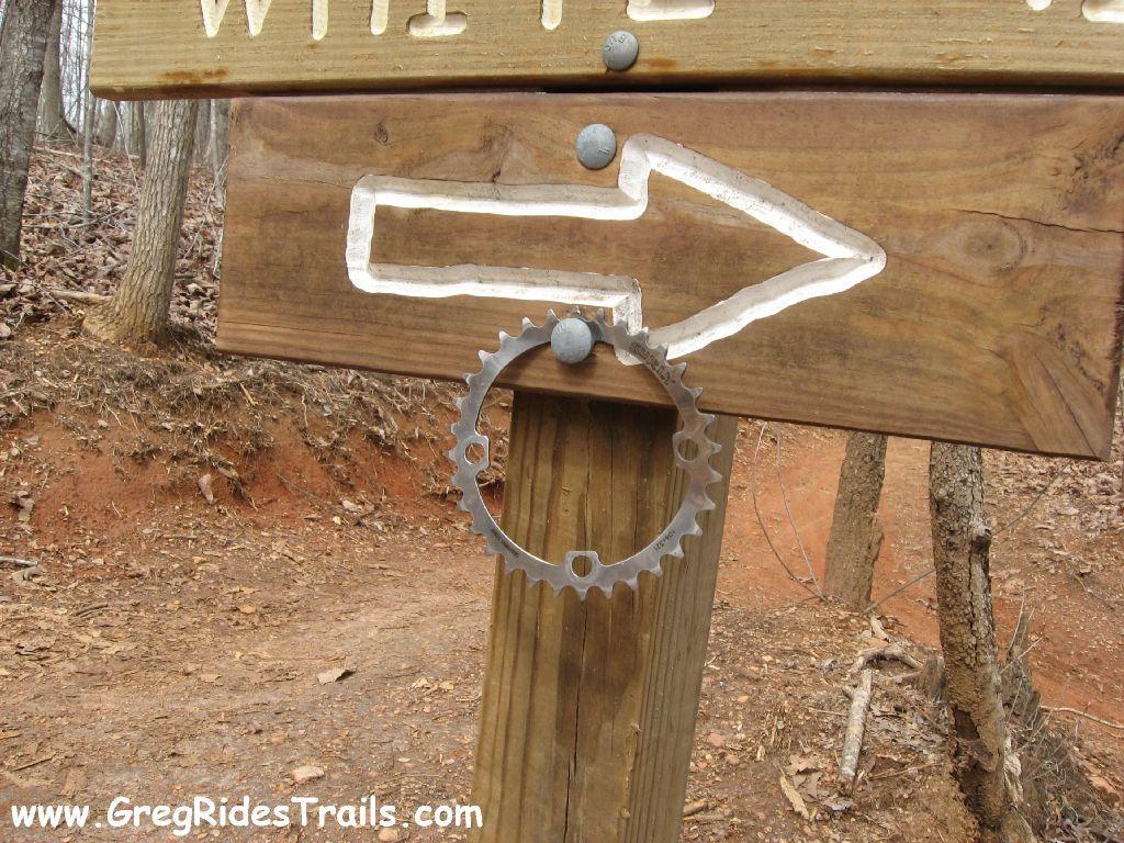 A wooden trail sign with an arrow pointing right, featuring a cut-out design and a metal bike chainring attached at the bottom. The background includes trees and a dirt path. Chicopee Woods mountain bike trail.