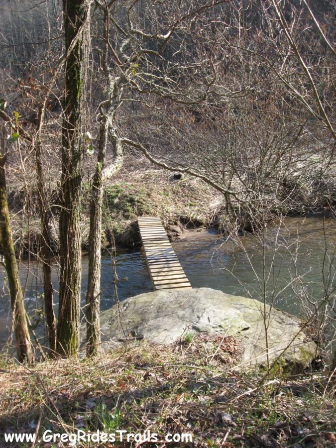 A wooden footbridge spanning a small stream, surrounded by bare trees and underbrush. A large rock is visible on one side of the bridge, with the water gently flowing beneath it. The scene captures a peaceful natural setting in early spring. Canada Creek mountain bike trail.