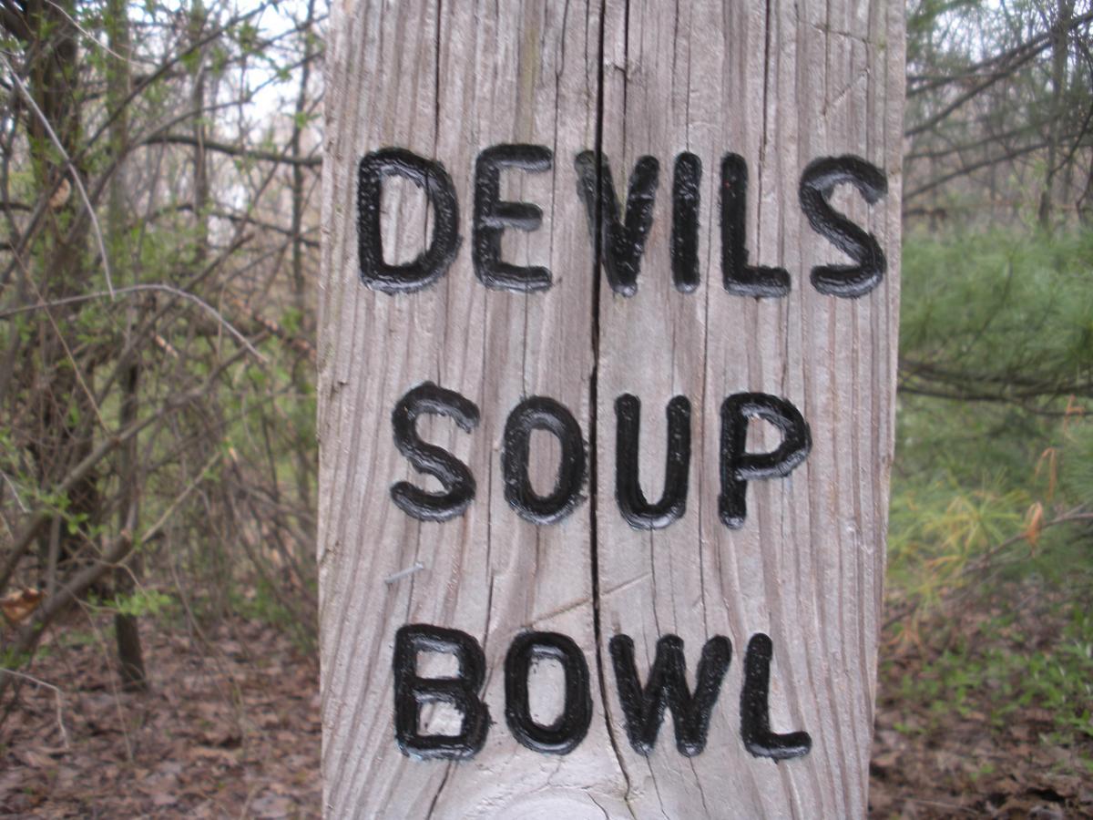 A wooden sign with the words "DEVILS SOUP BOWL" painted in bold black letters, set against a backdrop of trees and foliage in a natural outdoor setting. Yankee Springs mountain bike trail.