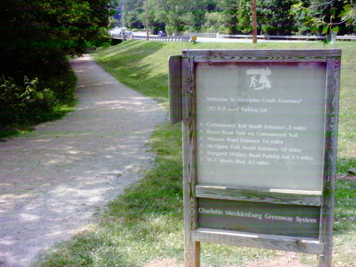 "Sign marking the entrance to McAlpine Creek Greenway, featuring directions and distances to various trails and parks, with a pathway leading through a wooded area." Mcalpine Park Greenway mountain bike trail.