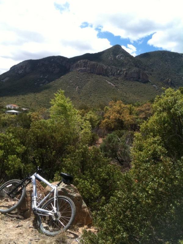 A mountain bike resting on a rock in a lush, green landscape, with mountains rising in the background under a partly cloudy sky. Brown Canyon mountain bike trail.