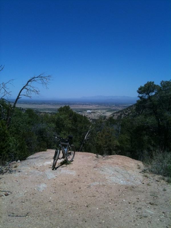 A mountain bike parked on a rocky outcrop overlooking a vast valley. The scene features clear blue skies, distant mountains, and scattered greenery in the foreground. Brown Canyon mountain bike trail.