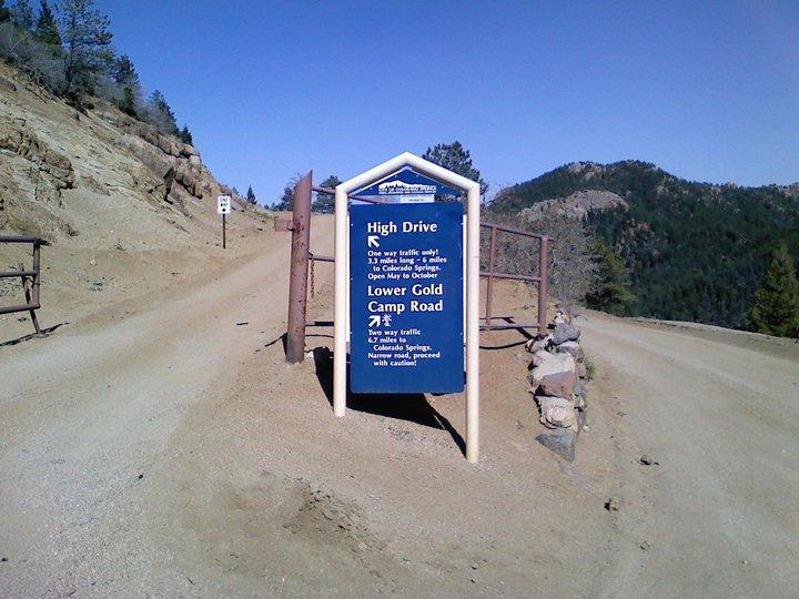 Signpost at the intersection of High Drive and Lower Gold Camp Road, detailing traffic regulations and directions. High Drive is designated for one-way traffic, while Lower Gold Camp Road allows two-way traffic, with caution advised for narrow sections. The surrounding landscape features rocky terrain and pine trees under a clear blue sky. Captain Jack's mountain bike trail.