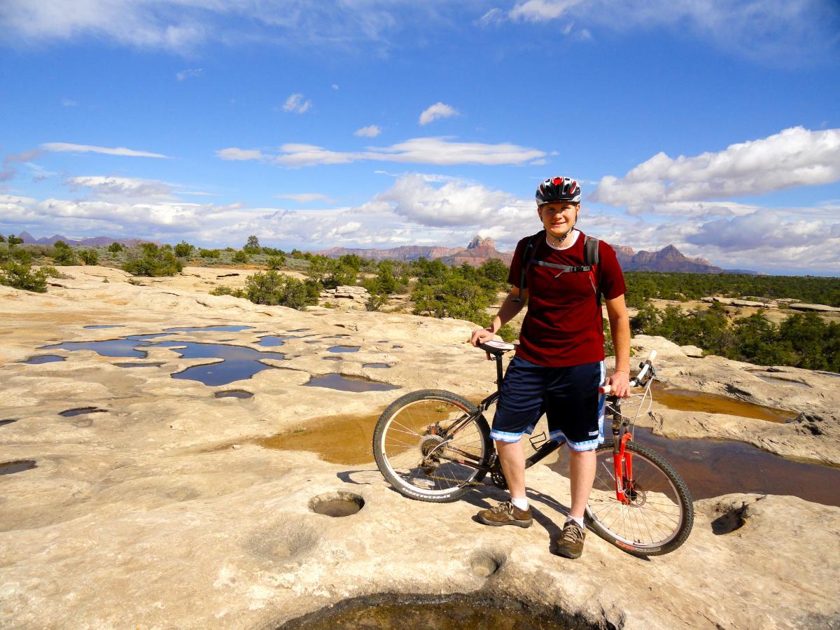 A person wearing a red helmet and a red shirt stands on rocky terrain holding a mountain bike. The landscape features patches of water, greenery in the background, and blue skies with scattered clouds. Gooseberry Mesa mountain bike trail.