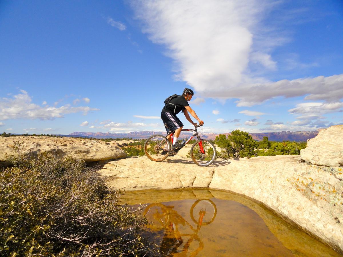 A mountain biker navigating a rocky terrain with a clear blue sky overhead. The rider is balancing on the edge of a rock formation, with a puddle reflecting the bike. Surrounding the area are patches of green vegetation and distant mountain ridges under a partly cloudy sky. Gooseberry Mesa mountain bike trail.