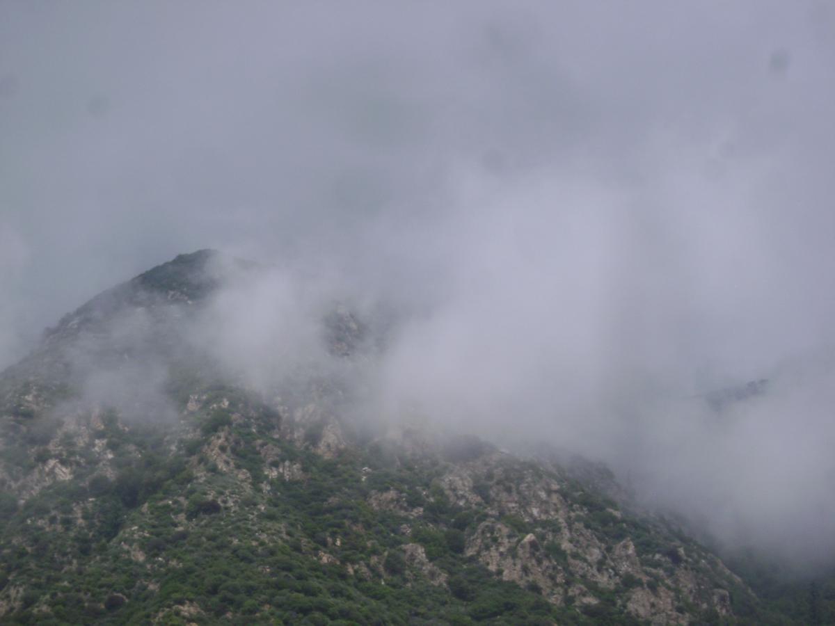 A mountain covered in fog, with lush greenery blending into rocky slopes. The sky above is gray and overcast, creating a misty atmosphere as clouds envelop the peak. Echo Mountain Loop mountain bike trail.