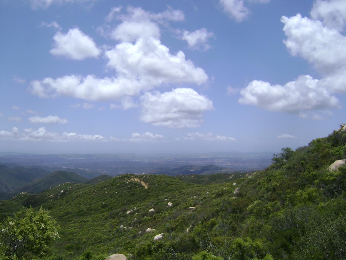 A panoramic view of rolling green hills under a bright blue sky with fluffy white clouds. The landscape features a mix of vegetation and rocky outcrops, with distant mountains visible in the background, creating a serene natural scene. San Juan Trail mountain bike trail.