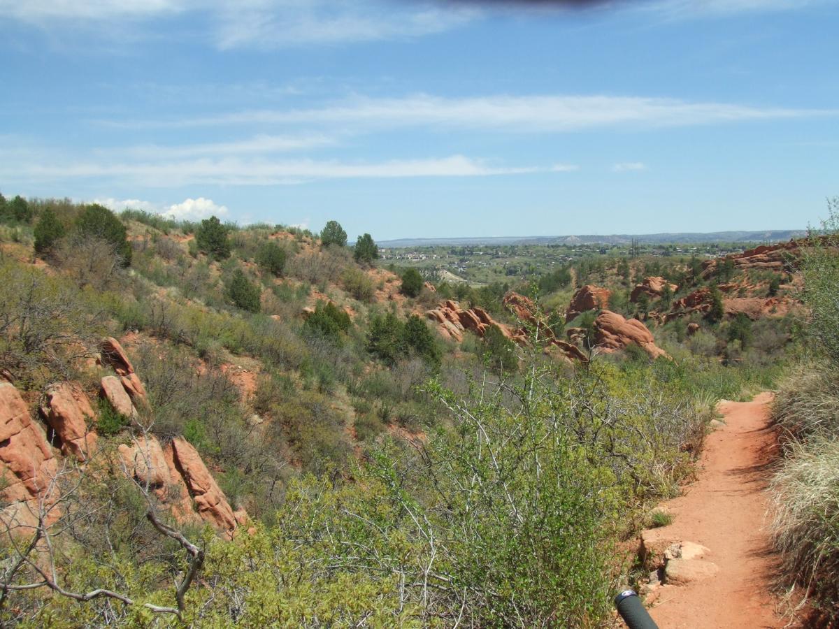 A winding dirt trail leads through a scenic landscape of rolling red rock formations and greenery under a clear blue sky. The terrain features bushes and small trees on the hillsides, with a view of distant hills and valley in the background. Red Rock Canyon mountain bike trail.