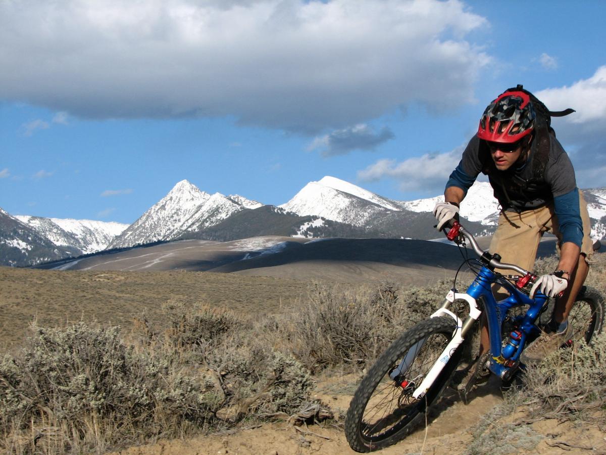 A mountain biker navigating a rocky trail with snow-capped mountains in the background under a partly cloudy sky. The cyclist is leaning forward on a blue mountain bike, wearing a helmet and protective gear, showcasing an adventurous outdoor scene. Discovery Hill Trails mountain bike trail.