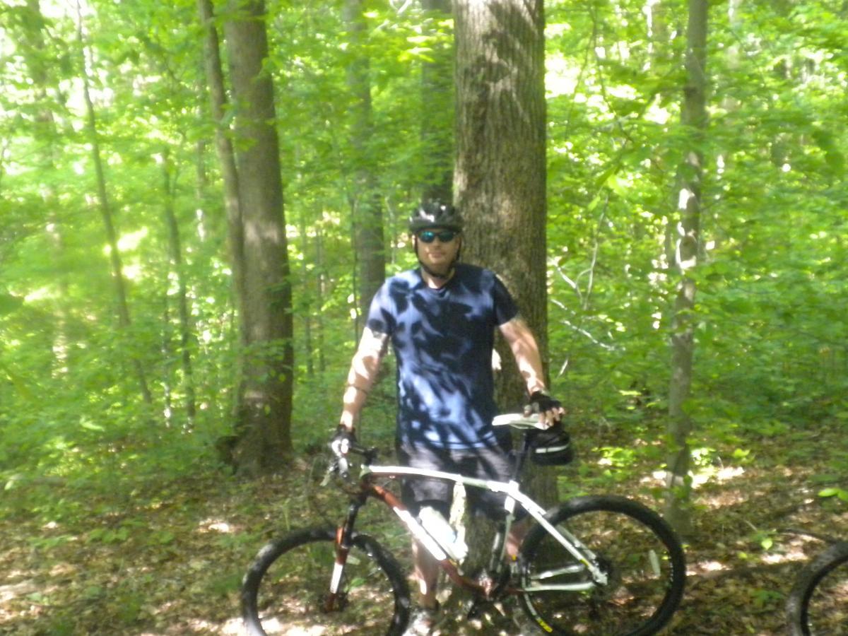 A person standing next to a mountain bike in a forested area, surrounded by green trees and sunlight filtering through the foliage. The individual is wearing a helmet and sunglasses, dressed in a navy blue shirt, and appears to be resting on a biking trail. Rosaryville State Park mountain bike trail.