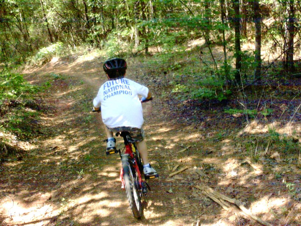 A child riding a bicycle on a winding dirt trail through a forest, wearing a t-shirt that reads "FUTURE NATIONAL CHAMPION." The scene features lush greenery and dappled sunlight filtering through the trees. Forks Area Trail System (FATS) mountain bike trail.