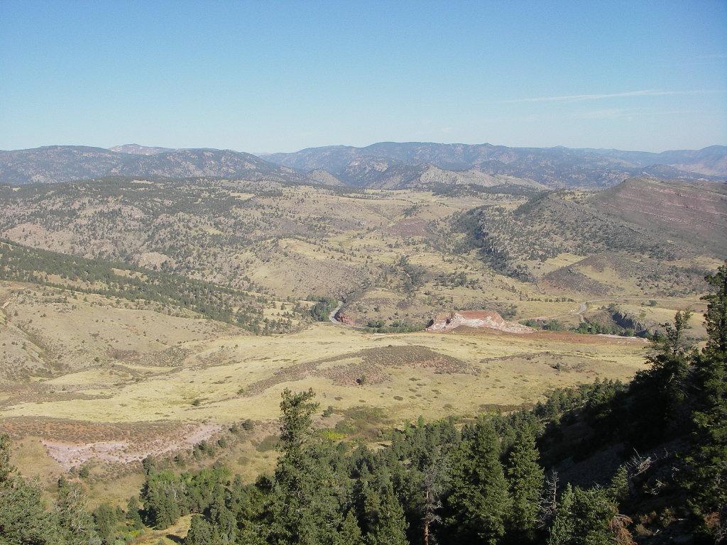 A panoramic view of a mountainous landscape featuring rolling hills, a winding river, and patches of trees. The scene captures a serene atmosphere under a clear blue sky, showcasing the natural beauty of the terrain. Heil Valley Ranch mountain bike trail.