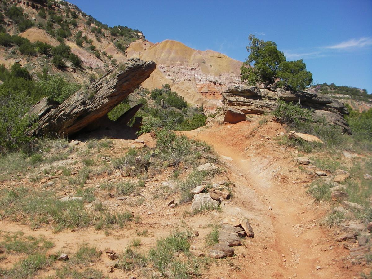 A rocky landscape featuring a dirt trail winding through green vegetation and large stone formations, with a backdrop of colorful hills under a clear blue sky. Palo Duro Canyon mountain bike trail.