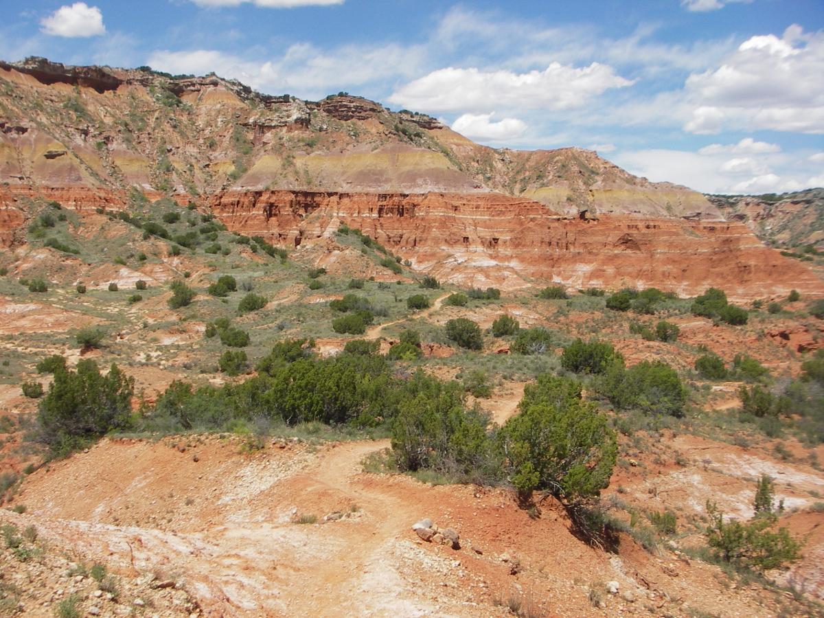A panoramic view of a rugged, red rock landscape with varying elevations, dotted with green shrubs and sparse vegetation under a blue sky with fluffy white clouds. The terrain features steep cliffs and gentle slopes, showcasing the natural beauty of the area. Palo Duro Canyon mountain bike trail.
