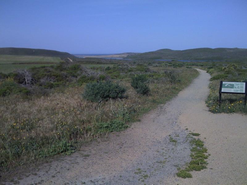 Pathway leading through a grassy area with shrubs, surrounded by rolling hills and a view of distant water. A sign is visible along the trail, providing information about the area. The sky is clear and blue, indicating a sunny day. Abbotts Lagoon mountain bike trail.