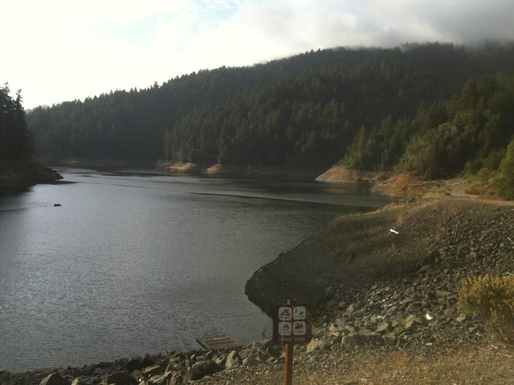 A serene lake surrounded by forested hills, with a rocky shoreline and a small boat on the water. A signpost with various icons is visible on the shore, indicating recreational activities. The sky is partially cloudy, and the atmosphere is calm and tranquil. Cross Marin Trail To Kent Lake mountain bike trail.