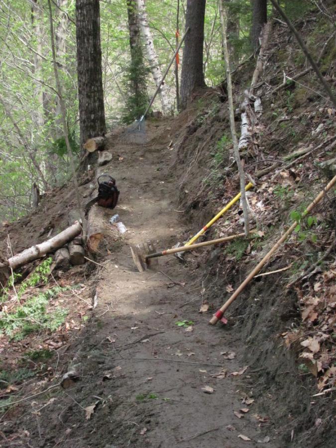 A narrow dirt trail running through a forest, flanked by trees. Various tools, including a rake and two digging implements, are positioned near the trail, alongside some logs and branches. A backpack is partially visible on the ground. The scene is well-lit by natural sunlight filtering through the leaves above. Franklin Falls mountain bike trail.