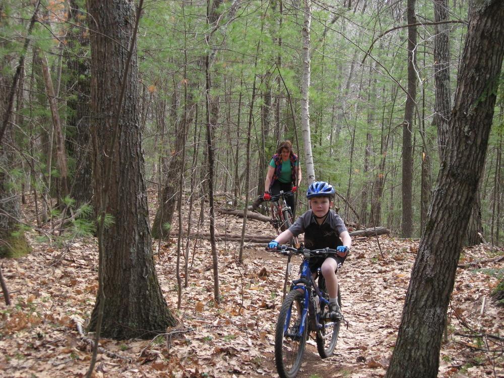 A young child riding a bicycle on a dirt trail in a forest, while an adult follows behind on another bike. The scene features tall trees, scattered leaves on the ground, and lush greenery surrounding the path. Franklin Falls mountain bike trail.