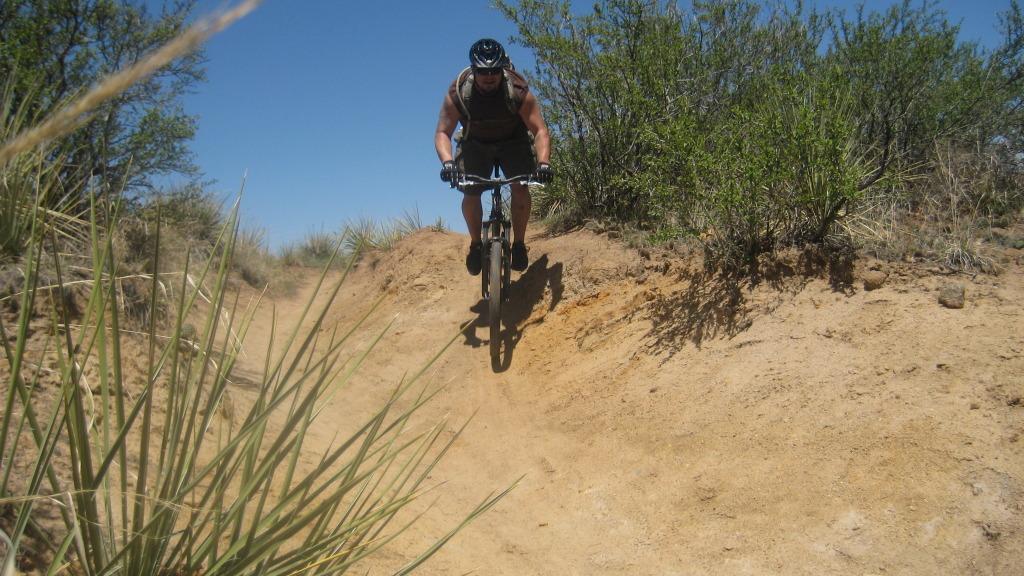 A person riding a mountain bike down a sandy, dirt trail, surrounded by sparse vegetation and blue sky. The rider is positioned mid-jump, showcasing an active outdoor scene. Palmer Park mountain bike trail.