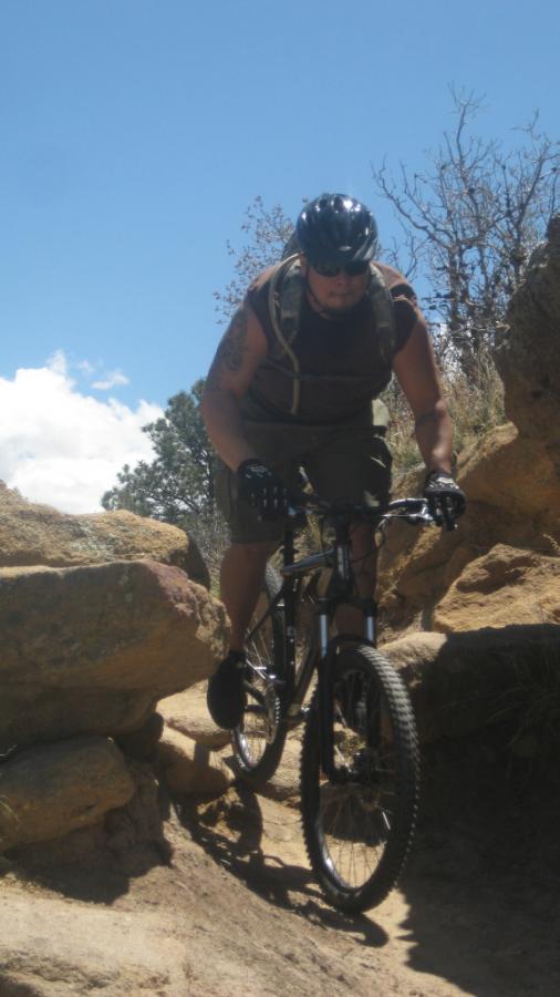 A mountain biker navigating a rocky trail under a clear blue sky, wearing a helmet and protective gear. The cyclist is focused and in motion, maneuvering through large boulders amidst a natural landscape. Palmer Park mountain bike trail.