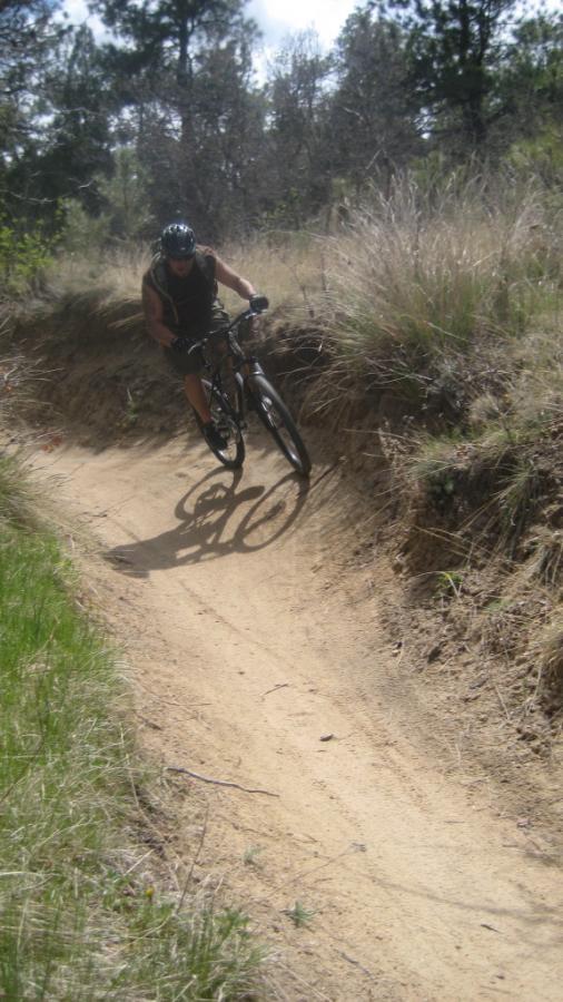 A person riding a mountain bike on a dirt trail surrounded by tall grass and trees, leaning into a turn. The scene captures a dynamic outdoor activity in a natural setting. Palmer Park mountain bike trail.