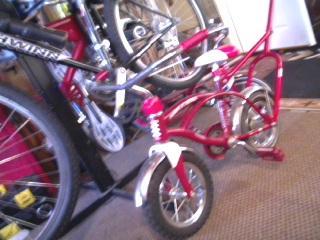 A red bicycle with smaller wheels parked next to larger bicycles in a bike shop or garage setting. The focus is on the distinct size and color of the smaller bike.