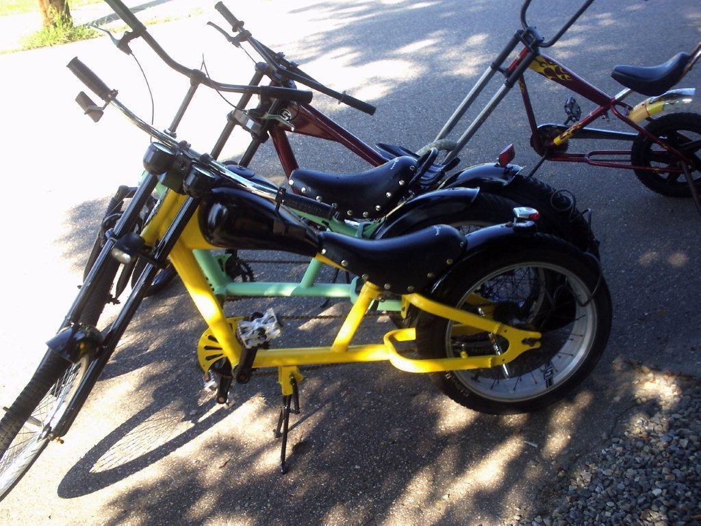 A close-up view of colorful custom bicycles parked on a paved surface, featuring a yellow and green two-seater bike with black seats, alongside other unique-style bikes. The setting is outdoors, with dappled sunlight filtering through the trees above.