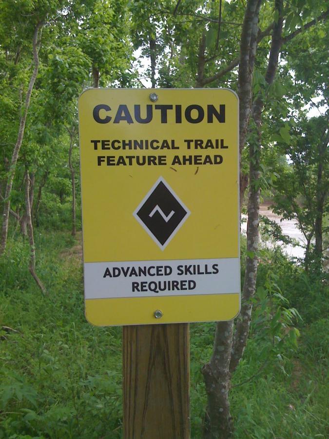 A yellow caution sign marking a technical trail feature ahead, indicating that advanced skills are required for navigation. The sign includes a black diamond shape with a zigzag symbol and is mounted on a wooden post, surrounded by green foliage and trees. Justin P. Brindley mountain bike trail.